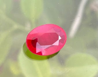 May include: A faceted, oval-shaped, deep red gemstone. The gem has a polished surface with multiple facets that reflect light. The stone is set against a blurred green and gray background, creating a contrast that highlights the gem's color and brilliance.