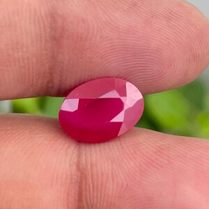 May include: A faceted, oval-shaped ruby gemstone. The ruby is a deep, vibrant red color with multiple facets that reflect light. The gemstone is held between two fingers, with a blurred green background.
