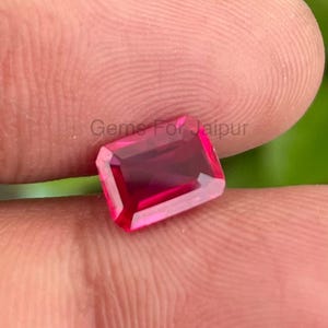 Può includere: Una gemma di rubino rettangolare e sfaccettata, di colore rosso intenso. La gemma è tenuta tra le dita, con il testo "Gems For Jaipur" visibile sullo sfondo. Il taglio della pietra ne evidenzia la chiarezza e la brillantezza.