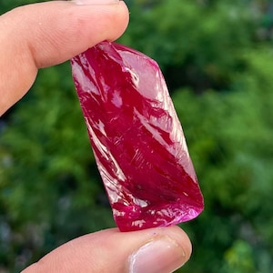 May include: A raw ruby gemstone, held between fingers. The ruby has a deep red colour with a slightly translucent quality. The surface has a textured, crystalline appearance, reflecting light. The background is a blurred green.