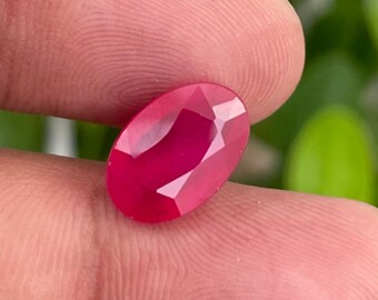 以下が含まれることがあります： ファセット加工された、楕円形のルビーの宝石。宝石は深く鮮やかな赤色で、光を捉えて反射する複数のファセットがあります。ルビーは指の間に挟まれ、背景はぼやけた緑色です。
