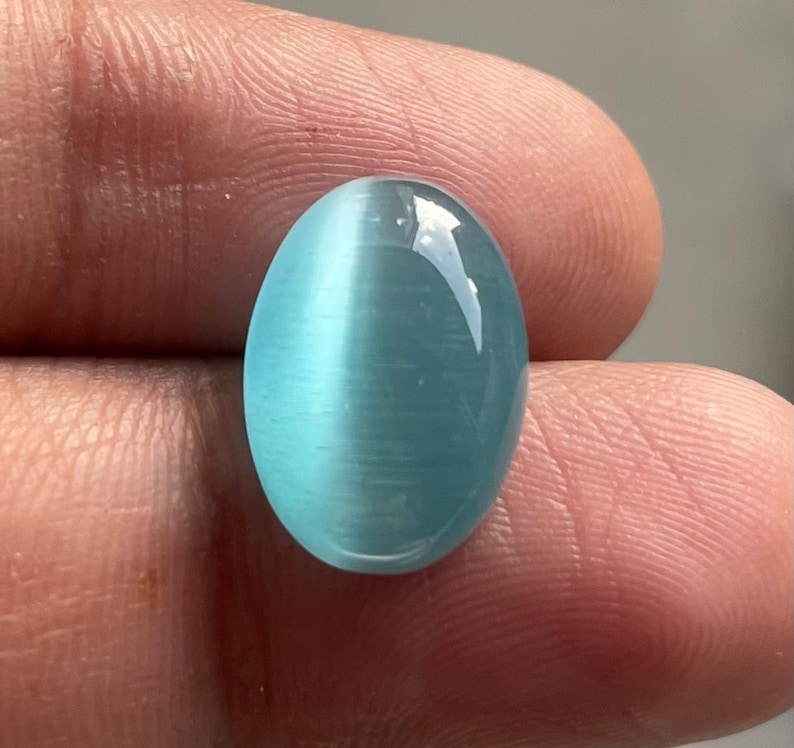 May include: A light blue oval cabochon gemstone with a chatoyant effect. The stone is smooth and polished.