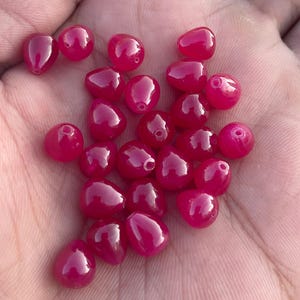 May include: A selection of glossy, deep pink beads in a teardrop shape. Each bead has a small hole for stringing. The beads are held in a hand, highlighting their smooth, reflective surfaces. Suitable for jewellery making.