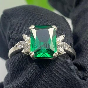 May include: A silver ring featuring a large, emerald-cut green gemstone as the centerpiece. Flanking the central stone are smaller, marquise-cut white gemstones. The ring is set against a dark background, highlighting the vibrant green of the main stone.