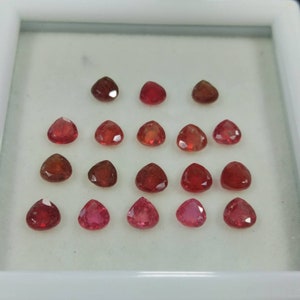 May include: A collection of 18 heart-shaped red gemstones displayed in a white box. The gemstones are arranged in three rows of six, with the largest gemstones in the back row and the smallest gemstones in the front row.