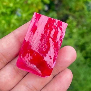 Puede incluir: Un cabujón rectangular que presenta un remolino vibrante de colores rojo brillante y rosa translúcido. La superficie es lisa y brillante, mostrando un patrón jaspeado con niveles variables de opacidad en toda la pieza, creando un contraste visual.