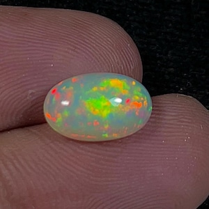 May include: An oval-shaped opal gemstone with a milky white base color. The opal displays flashes of red, orange, yellow, and green, creating a vibrant play-of-color effect. The gemstone is held between two fingers.