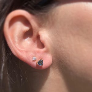 May include: A close-up of a person's ear with two earrings. The earring on the lower lobe is a small, triangular blue gemstone. The earring on the upper lobe is a small, round, clear gemstone.