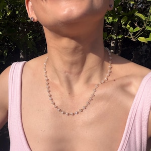 May include: A delicate gold chain necklace featuring small, translucent, light-colored beads spaced evenly along the length. The necklace rests against the neck, showcasing a simple link design that connects each individual bead in a continuous pattern.