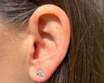 May include: A close-up of a person's ear with a small silver bee stud earring.