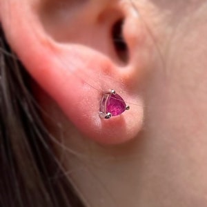 Pode incluir: Um close-up de um brinco de pedra preciosa rosa com forma triangular. O brinco é engastado em prata e é usado no lóbulo da orelha.