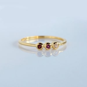 May include: A gold band ring with three small gemstones. The gemstones are a deep red, a lighter pink, and a clear white.