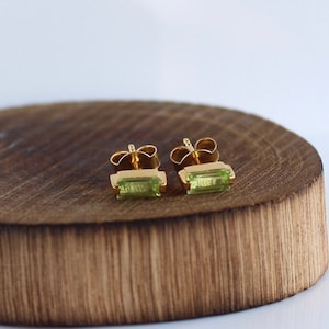 Può includere: Un paio di orecchini d'oro con pietre preziose rettangolari verdi. Gli orecchini sono su una superficie di legno.