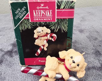 May include: A Hallmark Keepsake ornament featuring a tan puppy with a red collar, perched on a red and white candy cane. The ornament is from the New Collector's Series and comes in a decorative box.