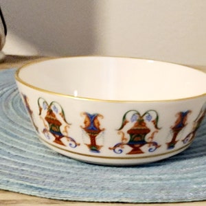 May include: A white ceramic bowl with a gold rim and decorative patterns in red, blue, and green. The bowl sits on a light blue placemat. The bowl's design features ornate, symmetrical motifs.