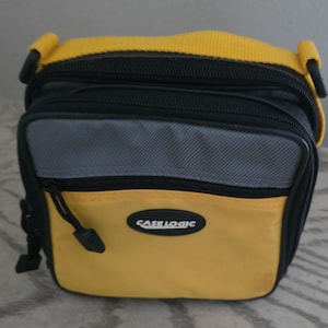 May include: A yellow and black Case Logic bag featuring a gray horizontal panel on the front, a zippered front pocket, and a yellow shoulder strap. The brand name Case Logic is printed in white text on a black oval patch on the front.