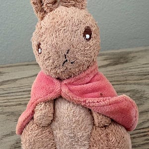 May include: A plush toy rabbit with a light brown body and long ears. The rabbit wears a pink cape around its neck. The toy is sitting on a wooden surface.