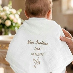 May include: A white muslin baby bib featuring embroidered text that reads Mon Bapt&ecirc;me, the name Noa, and the date 18.01.2025, accompanied by a small embroidered dove icon. The fabric has a soft, textured appearance with a triangular shape.