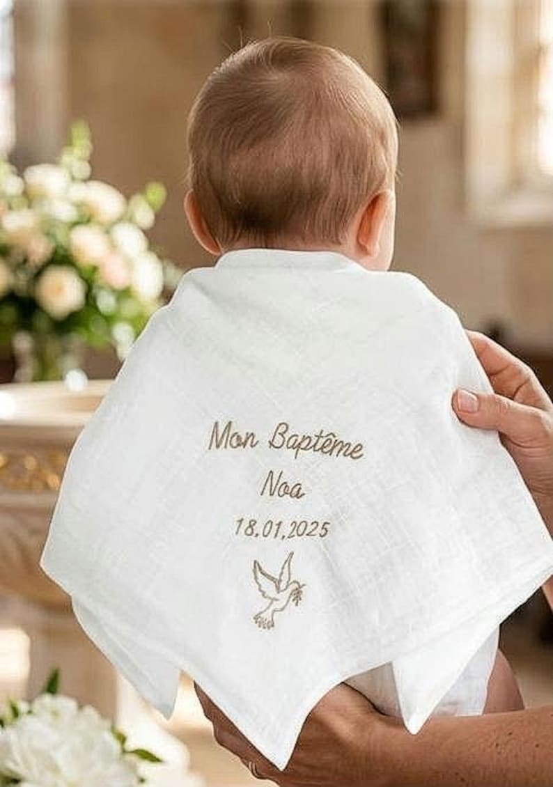 May include: A white muslin baby bib featuring embroidered text that reads Mon Bapt&ecirc;me, the name Noa, and the date 18.01.2025, accompanied by a small embroidered dove icon. The fabric has a soft, textured appearance with a triangular shape.