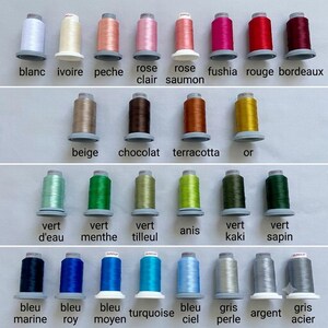 May include: A collection of twenty-seven spools of embroidery thread arranged in four rows, each labeled with its color name in French. The spools display a wide spectrum of colors including white, pinks, reds, browns, greens, blues, and grays.