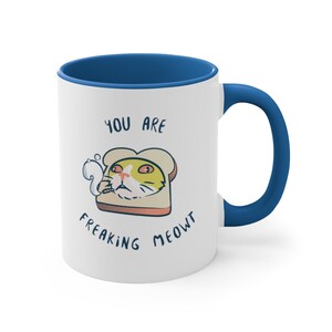May include: White ceramic mug with a blue rim and handle. The mug features a cartoon cat with a cigarette in its mouth, inside a slice of bread. The text "You are freaking meowt" is printed on the mug.