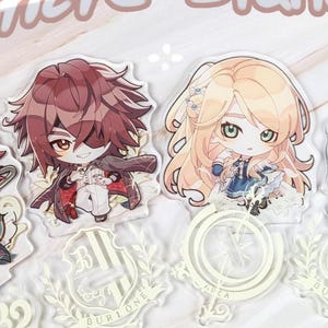 Könnte beinhalten: Vier Acrylständer mit Anime-Charakteren mit leuchtenden Haaren und Outfits. Jeder Ständer hat ein einzigartiges Design und wird von einem dekorativen Sockel getragen. Der Text "Pioriere Standees" ist in Kursivschrift geschrieben.