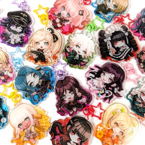 May include: A collection of acrylic keychains featuring various chibi-style anime characters with diverse hair colours and outfits. Each charm includes a colourful star-shaped plastic clasp and displays a unique character illustration with glitter accents.