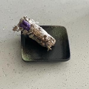 May include: A white sage smudge stick with purple flowers and dried herbs, tied with twine, rests on a small, square, black ceramic dish with a green rim. The dish sits on a speckled gray surface.