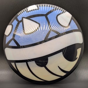 May include: A disc golf disc with a unique design. The disc features a light blue and white pattern with black accents. The text "2025 TOUR SERIES" is printed on the disc. The disc is round and appears to be made of plastic.