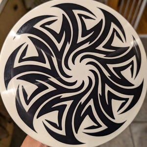 Pode incluir: Um disco branco com um design tribal preto. O design é um padrão de redemoinho de ângulos agudos e curvas.