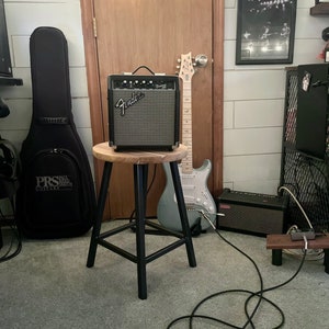 May include: A black Fender guitar amplifier sits on a wooden stool with a black metal frame. A blue electric guitar is leaning against the stool. A black guitar case is on the floor to the left of the stool. A black cable is coiled on the floor.