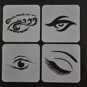 Puede incluir: Cuatro plantillas cuadradas con diseños de ojos. Cada plantilla presenta un diseño de ojo negro diferente, incluyendo pestañas detalladas y varias formas. Las plantillas están sobre una superficie de madera oscura, ideales para maquillaje o proyectos artísticos.