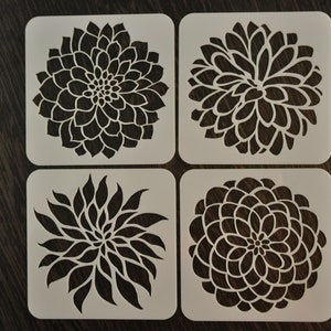 May include: Set of four white plastic stencils featuring different floral designs. Each stencil is square shaped and features a black silhouette of a flower.