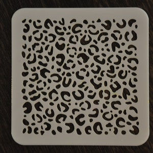 Pode incluir: Um estêncil de plástico branco com um design de estampa de leopardo. O estêncil é quadrado e tem uma superfície lisa.