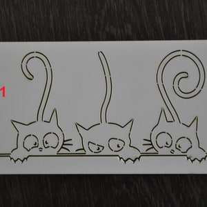 May include: White plastic stencil with three black cats peeking over a line. The cats have curled tails and simple facial features. The number "1" is printed in the top left corner.