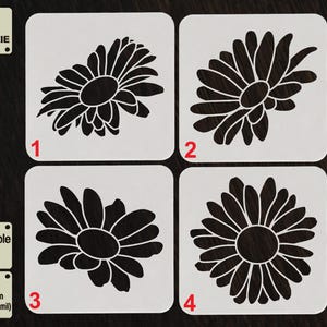 Puede incluir: Cuatro plantillas cuadradas blancas con diseños de margaritas negras, numeradas del 1 al 4. Las plantillas están etiquetadas como "TOOL FOR COOKIE", "Reusable Stencil" y "APET 1000 micras (1 mm / 40 mil)".