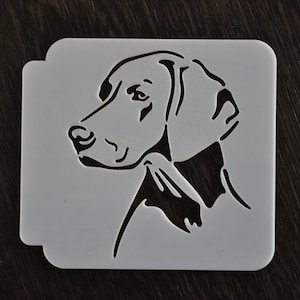 Può includere: Uno stencil di plastica bianca con la testa di un cane, il cane è rivolto a sinistra, lo stencil è su una superficie di legno scuro.