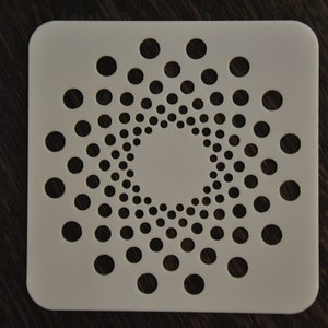 Könnte beinhalten: Weiße quadratische Schablone mit einem kreisförmigen Muster aus gleichmäßig verteilten Löchern. Die Löcher sind in konzentrischen Kreisen angeordnet und bilden ein radiales Design.
