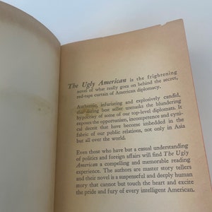 May include: Open book with text from "The Ugly American" novel. The text describes the novel's themes of American diplomacy, hypocrisy, and the blunders of top-level diplomats. The book's pages are aged with a light orange hue.