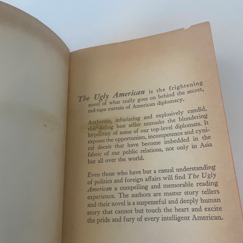 May include: Open book with text from "The Ugly American" novel. The text describes the novel's themes of American diplomacy, hypocrisy, and the blunders of top-level diplomats. The book's pages are aged with a light orange hue.