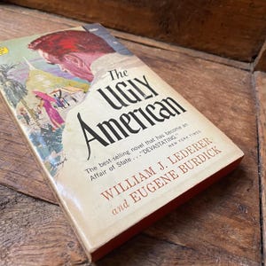 May include: A vintage copy of "The Ugly American" novel. The book cover features an illustration of a man's head and a colorful scene. The title is in large black letters, with the author's names below.