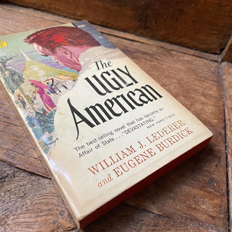May include: A vintage copy of "The Ugly American" novel. The book cover features an illustration of a man's head and a colorful scene. The title is in large black letters, with the author's names below.