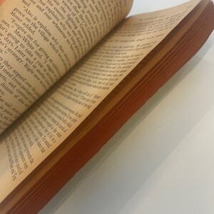May include: An open book with aged, yellowed pages and a reddish-brown edge. The text is in black, with the pages slightly curved. The book is angled, showing the text and the side of the pages.