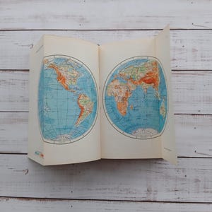May include: An open book displaying two circular world maps on its pages. The left page shows the Americas, while the right page depicts Europe, Africa, and Asia. The maps feature blue oceans and orange landmasses with thin black grid lines.