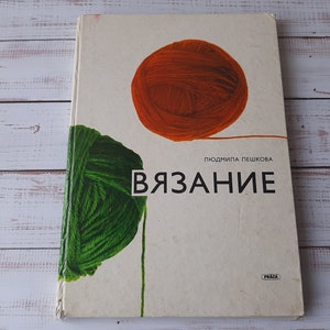 Könnte beinhalten: Ein rechteckiges Buch mit cremefarbenem Einband, das oben ein großes orangefarbenes Wollknäuel und auf der linken Seite ein grünes Wollknäuel zeigt. Der Text "ЛЮДМИЛА ПЕШКОВА" und "ВЯЗАНИЕ" ist schwarz gedruckt, mit einem kleinen "PRÁCA"-Logo unten rechts.