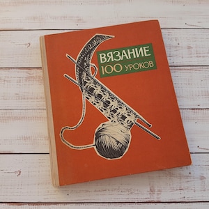 Könnte beinhalten: Ein gebundenes Buch mit einem rostorangen Einband, das eine grafische Illustration eines Wollknäuels und Stricknadeln zeigt. Der Text auf dem Einband lautet "ВЯЗАНИЕ 100 УРОКОВ" in weißen und gelben Buchstaben vor einem dunkelgrünen rechteckigen Hintergrund.