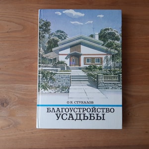 May include: A hardcover book with a white cover featuring a color illustration of a modern house with a stone wall and garden. The text O.K. STUKALOV and BLAGOUSTROISTVO USADBY is printed in black on the lower portion of the front cover.