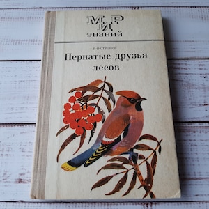 Puede incluir: Libro ruso vintage titulado "Amigos alados de los bosques". La portada presenta una ilustración de un pájaro posado en una rama con bayas rojas. El libro tiene una cubierta de color crema.