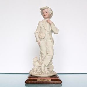 Op de afbeelding: Een crèmekleurig porseleinen beeldje van een jonge persoon in een pak, die zijn stropdas rechtzet. Een klein konijn zit aan hun voeten. Het beeldje is gemonteerd op een houten voet met de naam "N. Belcari".
