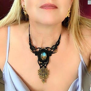 Peut inclure: Un collier en macramé noir avec une pierre de labradorite ovale centrale aux reflets bleus et dorés irisés. Le design comprend un tressage complexe en cordon noir, de petites perles métalliques et un pendentif en filigrane doré.